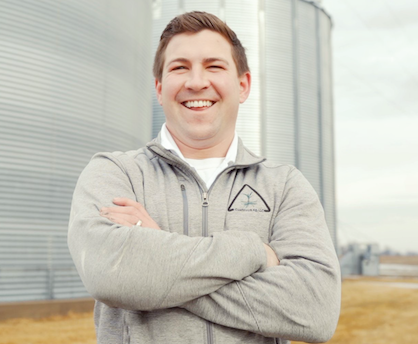 Mitchell Hora standing in a field in front of grain storage containers with his arms crossed.